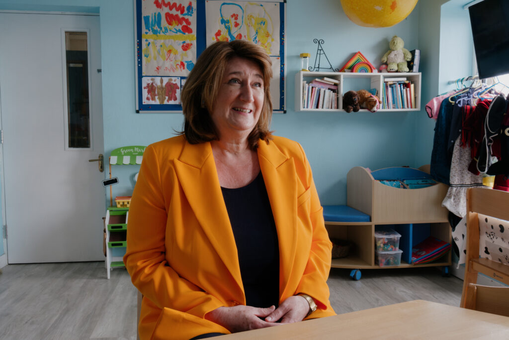 A woman sits in a classroom painted in bright colors wearing a bright orange jacket.