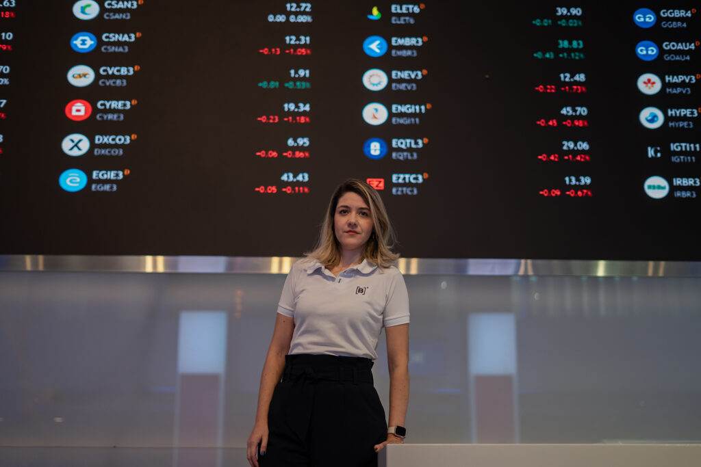 Christianne Bariquelli standing in front of a giant black screen displaying ticker symbols in vivid colors.