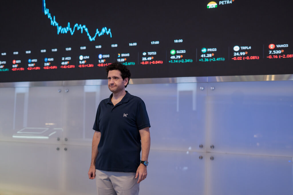 Marcos Albino Rodrigues standing in front of a huge black screen showing ticker symbols and graphs in vivid colors.