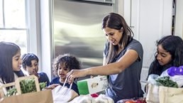 A mother is putting away groceries in the kitchen while her four children observe with curiosity. She has a relaxed expression as she inspects a box of crackers.