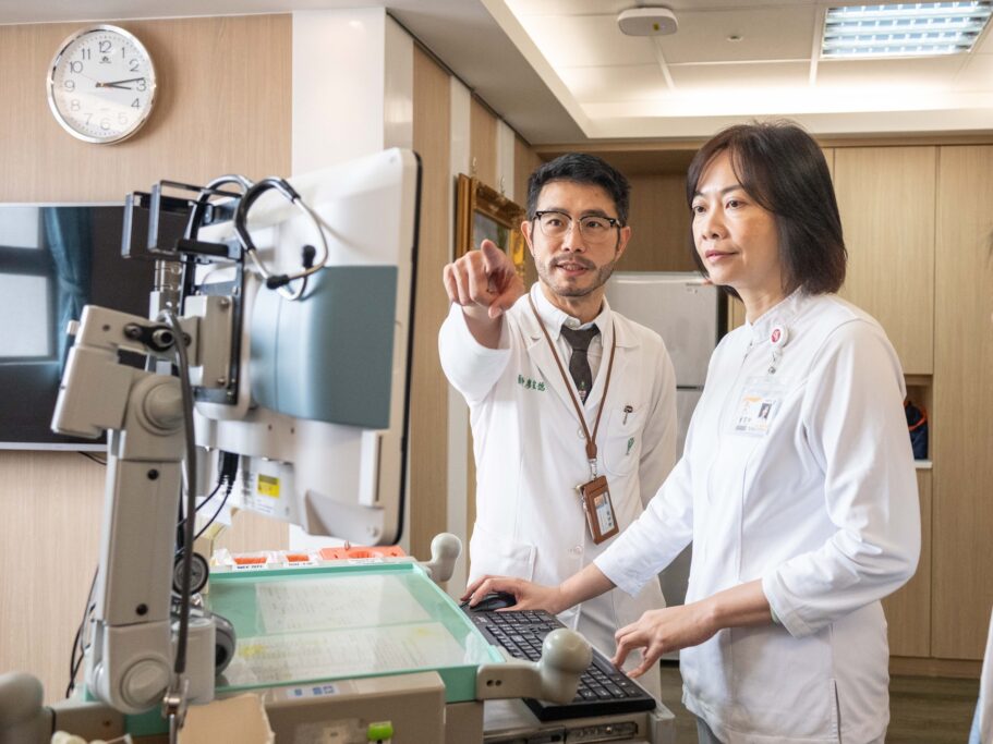 Two people in white lab coats standing in front of a computer in a hospital room