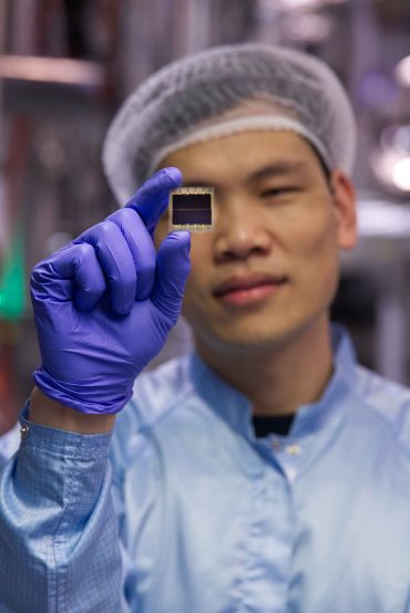 An researcher of Asian descent wearing a lab coat, hairnet, and latex gloves, holds up a wafer-thin material that looks like a computer chip.