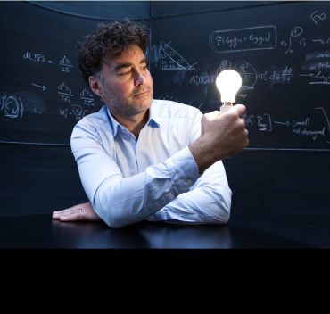 A white man with dark curly hair holds up an illuminated light bulb. He is sitting in front of a blackboard with mathematical equations written in chalk.