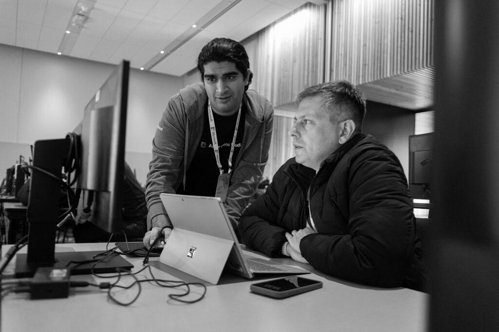 Two people sit at a conference looking at a computer screen and a tablet.