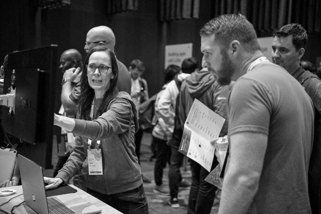 Two people at a conference look at a computer screen while a presenter points at it.