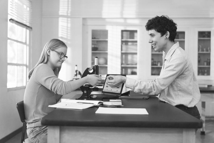 Decorative. A teacher hands a microscope slide to a student. The student is working with a microscope and a laptop at a table in a science classroom.