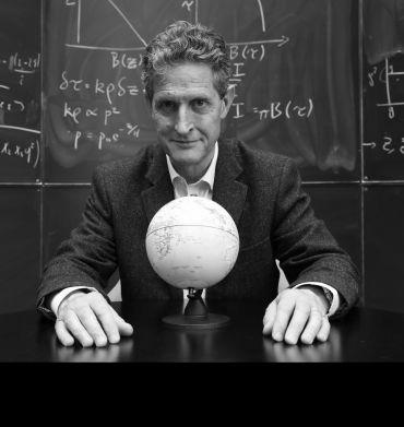 A portrait photograph of a white man wearing a dark suit, seated at the table with a 3D Globe model in front of him. In the background is a blackboard with mathematical equations scribbled on it.