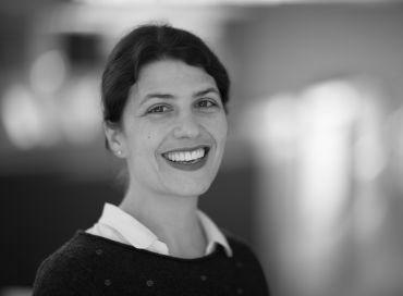 Portrait photograph of Professor Elena Seiradake, a white lady with dark hair standing against an out-of-focus interior backdrop.