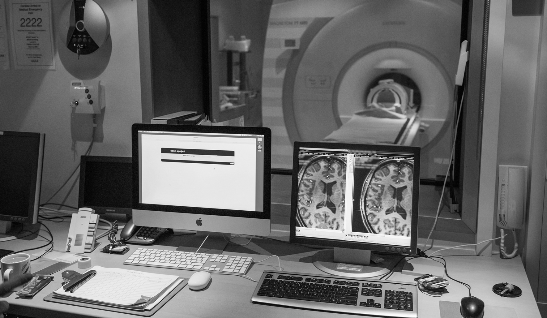 A medical imaging control room featuring two computer monitors displaying brain scan images, a keyboard, and various equipment, with a view of an MRI machine through a large observation window.