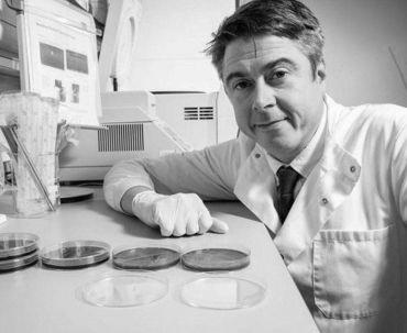 Professor Sam Shepphard: a white man in a laboratory wearing a lab coat. On the bench in front of him are petri dishes filled with coloured and clear agar media.