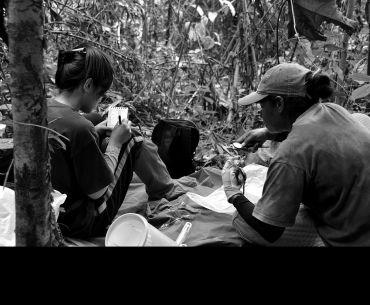 Two researchers sit on a tarp in a dense tropical forest. One writes in a notepad, while the other uses callipers to measure a small mammal. Surrounding them are tools, a bucket, and a backpack, indicating fieldwork in biodiversity research.