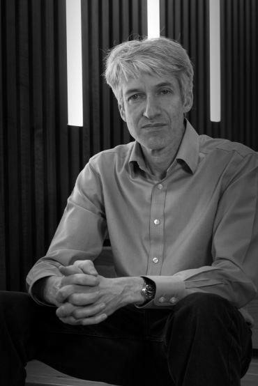 Professor David Lucas, a white man wearing a blue shirt and black trousers. He sits in a building with vertical strip lights in the background.