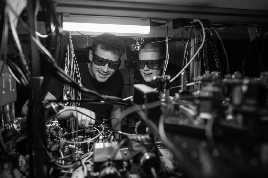 A male and female researcher wearing protective glasses look inside a quantum computer, made up of many wires and electrical components.