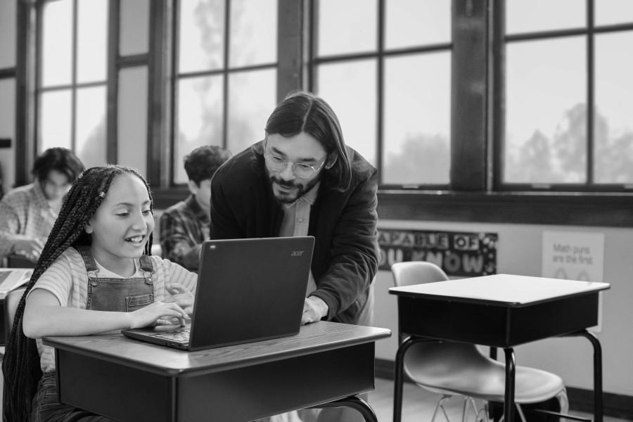 An educator in a classroom helps a student with schoolwork on her laptop.