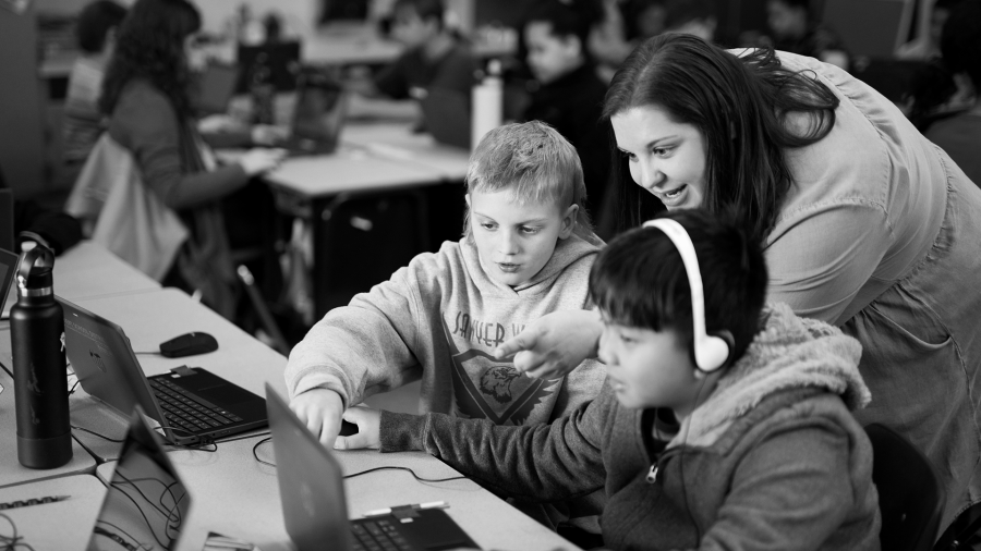 A teacher leans over two students at a desk helping them with Minecraft Education.