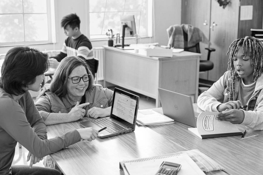 A classroom scene with students at communal desks and an educator leans in to help one of the students.